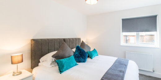 A neatly designed bedroom featuring a large bed with decorative pillows, a bedside lamp, and a window with a roller shade. The color palette includes light walls, gray bed linens, and teal accents.