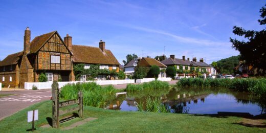 A picturesque village scene featuring traditional thatched-roof houses alongside a tranquil pond, surrounded by lush greenery and blue skies. The area conveys a serene countryside ambiance, inviting visitors to appreciate the idyllic setting.