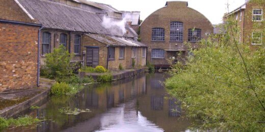 A tranquil scene depicting industrial buildings along a waterway, with lush greenery. The architecture features a distinctive rounded structure, and some buildings show smoke rising from their rooftops, set against a cloudy sky.