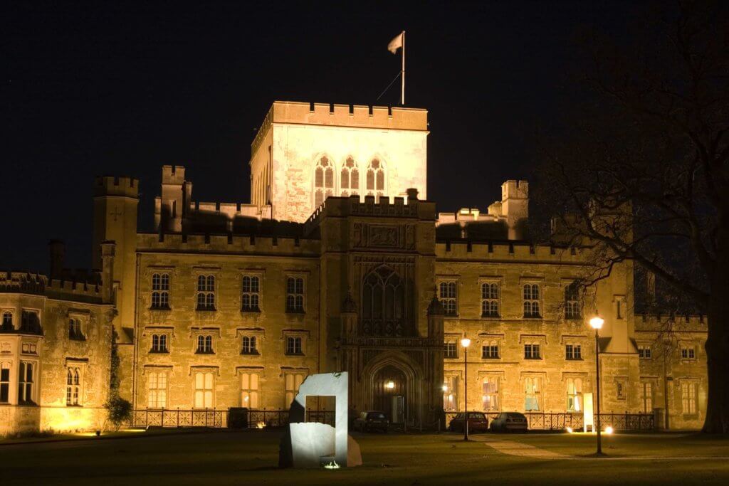 A grand historical castle illuminated at night, featuring turrets and a flag atop the central tower. The entrance is marked by an ornate archway, while a modern sculpture stands in the foreground, contrasting the traditional architecture.