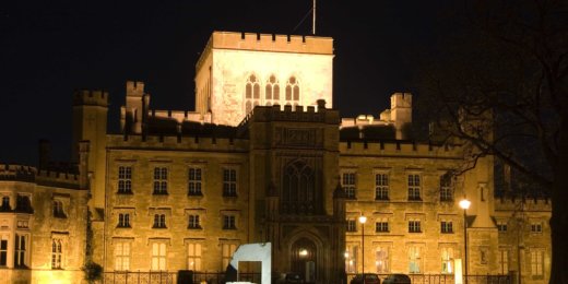 A grand historical castle illuminated at night, featuring turrets and a flag atop the central tower. The entrance is marked by an ornate archway, while a modern sculpture stands in the foreground, contrasting the traditional architecture.