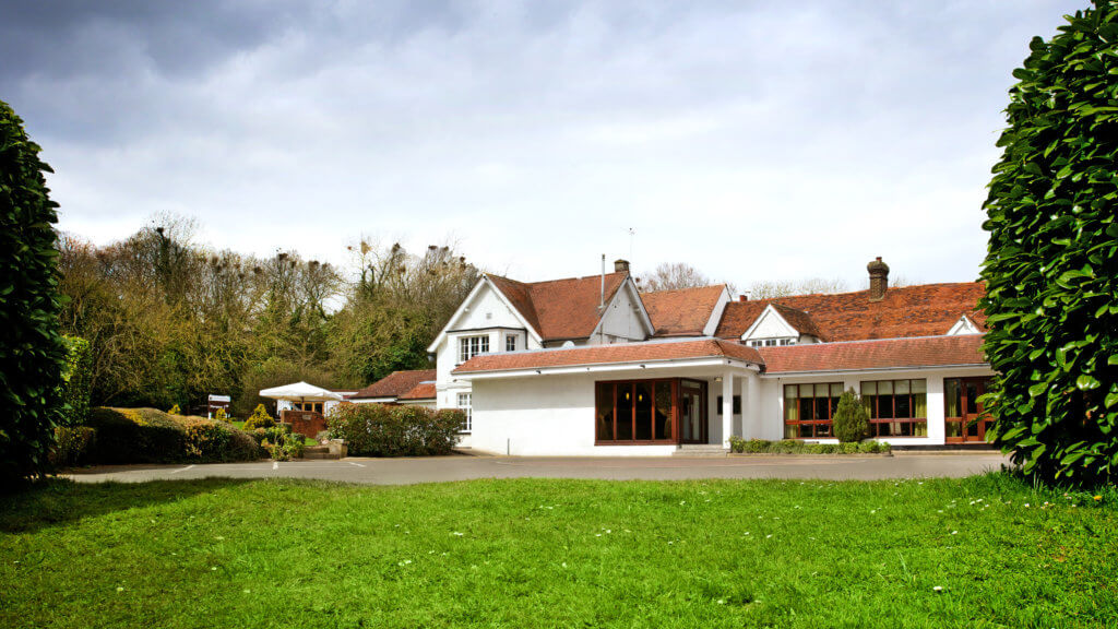 A modern, two-story house with a sloped roof and large windows sits on a green lawn, surrounded by lush trees and shrubs under a cloudy sky. The entrance features multiple wooden doors leading into the home.
