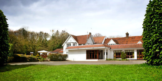 A modern, two-story house with a sloped roof and large windows sits on a green lawn, surrounded by lush trees and shrubs under a cloudy sky. The entrance features multiple wooden doors leading into the home.