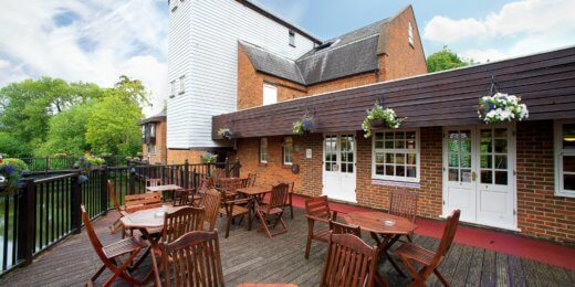 A scenic outdoor seating area featuring wooden tables and chairs, surrounded by greenery, with a two-story brick building in the background. The sky is partly cloudy, creating a pleasant atmosphere for relaxation.