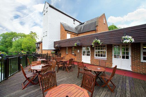 A wooden deck with tables and chairs overlooks a calm waterway, surrounded by greenery. The building features a mix of brick and wood paneling, with large windows and hanging flower baskets, set against a partly cloudy sky.