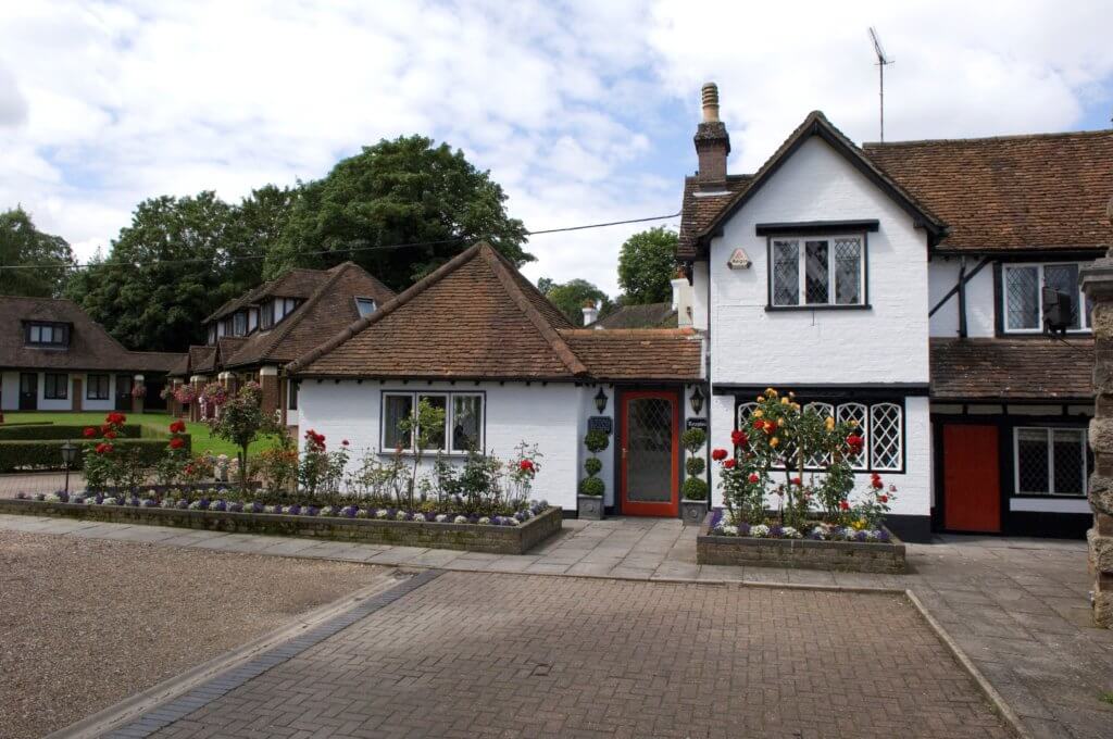 A charming two-story house with a thatched roof and white walls, surrounded by a flower garden. The front features decorative windows and a red door, set in a picturesque neighborhood with trees in the background.