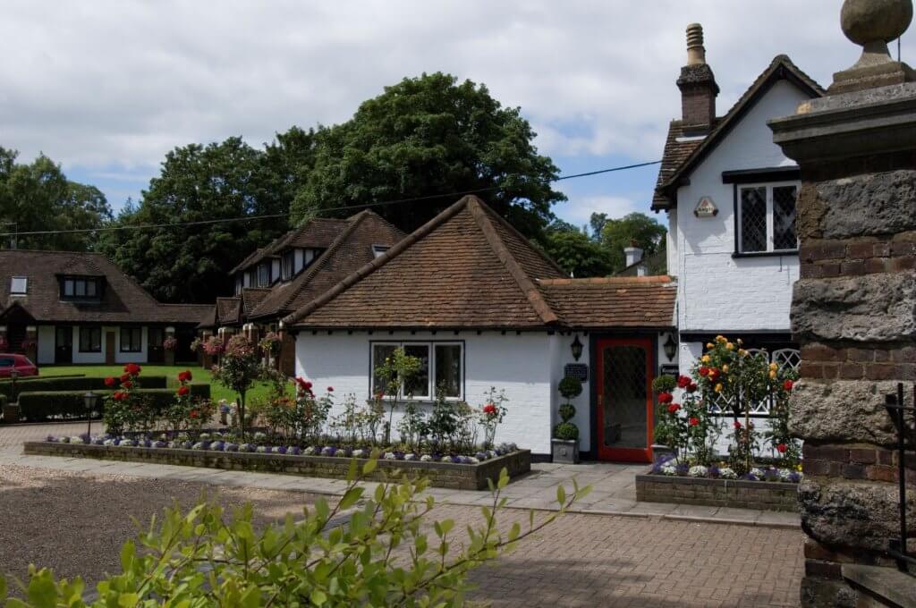 A charming cottage with a thatched roof and white exterior, surrounded by lush greenery and colorful flowerbeds. The entrance features a red door, adding a welcoming touch to the picturesque setting.