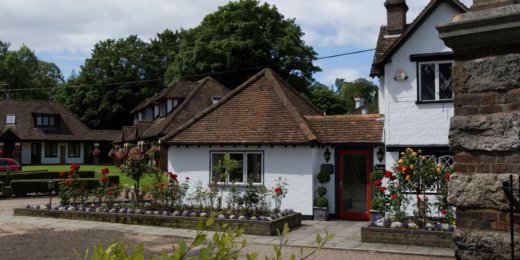 A charming cottage with a thatched roof and white exterior, surrounded by lush greenery and colorful flowerbeds. The entrance features a red door, adding a welcoming touch to the picturesque setting.