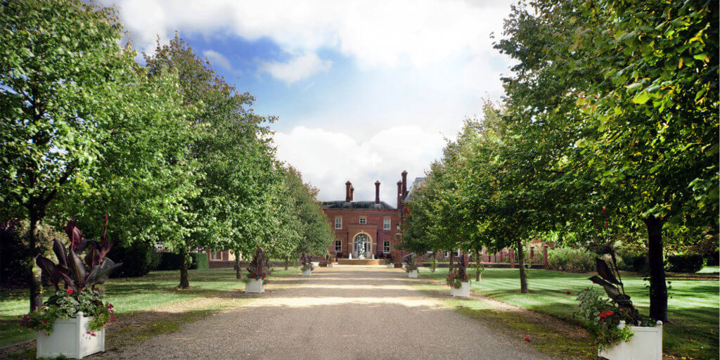 A wide gravel pathway leads towards an elegant brick mansion, framed by lush green trees and decorative planters. The sky above is partly cloudy, creating a serene and inviting atmosphere.