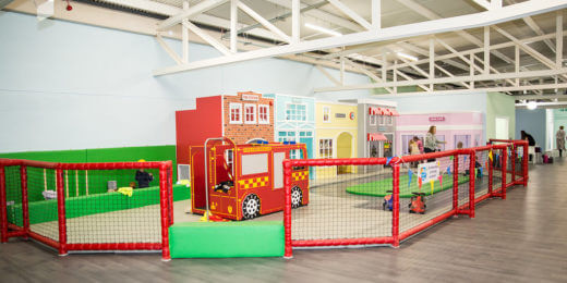 A colorful indoor play area features a fenced section with a toy fire truck and themed buildings, encouraging imaginative play for children. Brightly painted structures and a green play surface enhance the vibrant atmosphere.