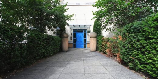 A pathway leads to a blue door framed by two large stone planters. Lush greenery and hedges line the sides, creating a serene and inviting entrance.