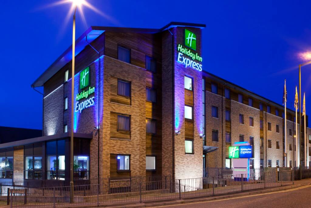 Holiday Inn Express hotel exterior illuminated at night, featuring a modern design with large windows and colorful signage. The building is surrounded by a parking area and streetlights, creating a welcoming atmosphere.