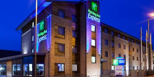 Holiday Inn Express hotel exterior illuminated at night, featuring a modern design with large windows and colorful signage. The building is surrounded by a parking area and streetlights, creating a welcoming atmosphere.