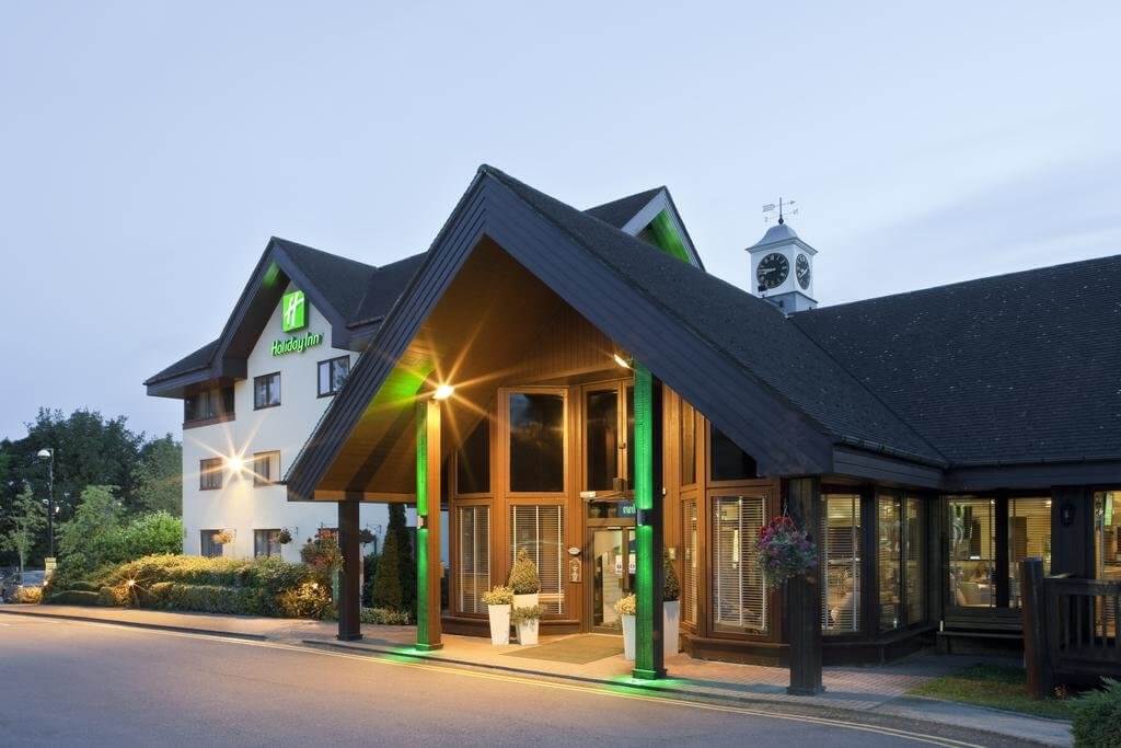 The image shows the entrance of a modern hotel with a welcoming wooden facade, illuminated by outdoor lights. A clock tower is visible in the background under a twilight sky, creating a cozy atmosphere. The entrance features large glass doors and decorative planters.