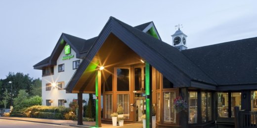 The image shows the entrance of a modern hotel with a welcoming wooden facade, illuminated by outdoor lights. A clock tower is visible in the background under a twilight sky, creating a cozy atmosphere. The entrance features large glass doors and decorative planters.