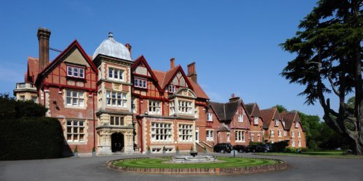 A large, picturesque red-brick building with ornate architectural features, surrounded by greenery and a circular driveway. The sky is clear and blue, enhancing the charming appearance of the property.