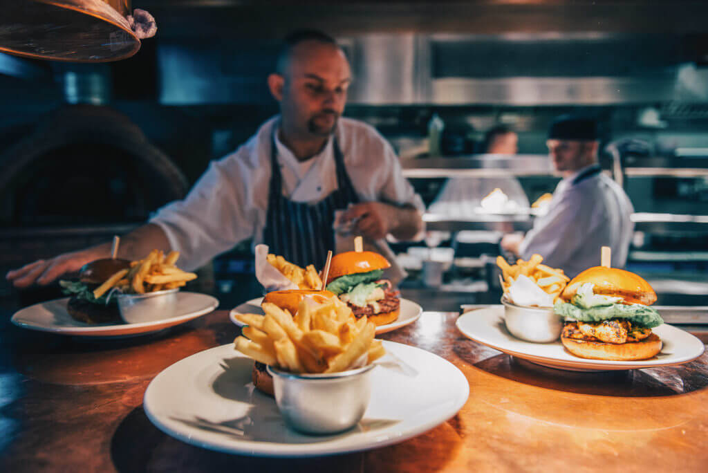 A chef serves three plates of burgers and fries in a busy kitchen, highlighting the vibrant atmosphere of culinary preparation.