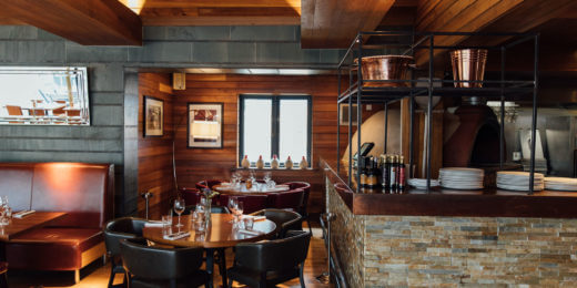 A cozy restaurant interior featuring wooden walls, a stone bar area, and elegant dining tables with dark chairs. Natural light streams through a window, creating a warm atmosphere. Copper decor elements add a touch of sophistication to the inviting space.