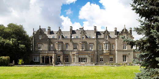 A grand, historic mansion with intricate architecture stands prominently on a lush green lawn, surrounded by trees under a bright blue sky with fluffy white clouds.