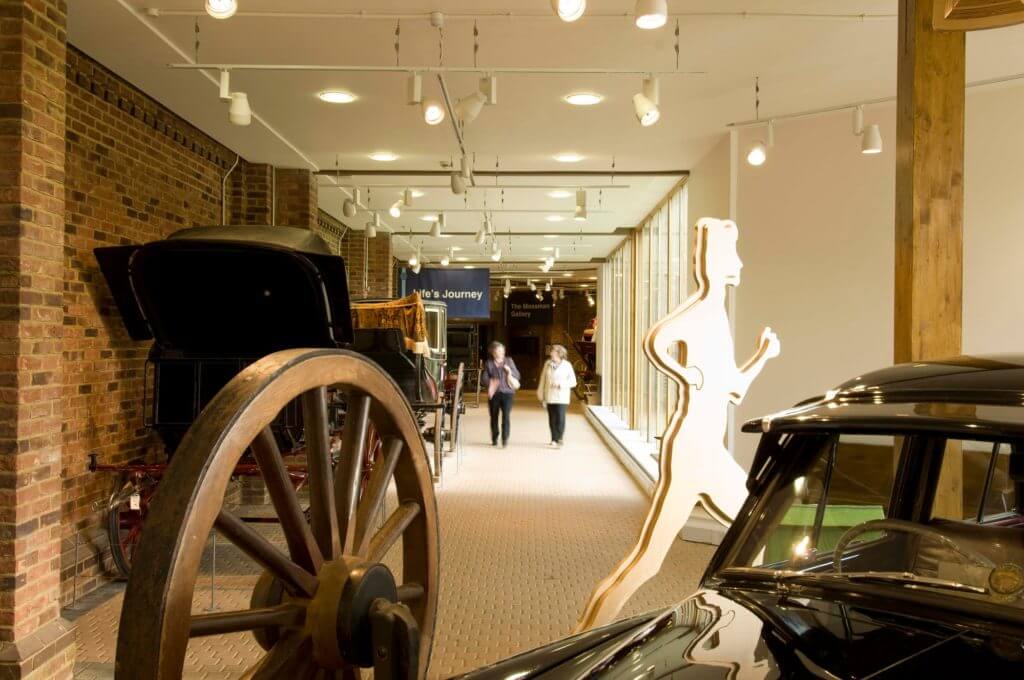 The image shows an indoor exhibit featuring vintage vehicles with a walking figure silhouette in the foreground. Two people are seen walking in the background along a well-lit corridor lined with brick walls and large windows.