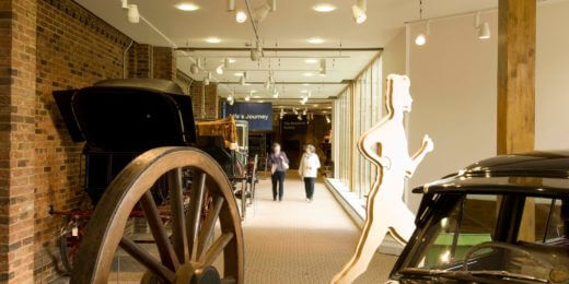 The image shows an indoor exhibit featuring vintage vehicles with a walking figure silhouette in the foreground. Two people are seen walking in the background along a well-lit corridor lined with brick walls and large windows.