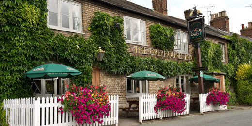 A charming pub with ivy-covered stone walls, green umbrellas over outdoor tables, and vibrant flower arrangements in front. The welcoming facade invites patrons to enjoy the outdoor seating in a picturesque setting.