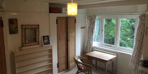 A cozy bedroom featuring a wooden chair, a small table, a shelf, and a window with curtains that lets in natural light. The walls are adorned with simple decor, and the room has a warm, inviting atmosphere.