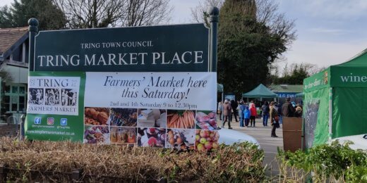 A sign for Tring Market Place displays information about a farmer's market scheduled for Saturday, with people gathered in the background and various market stalls visible.