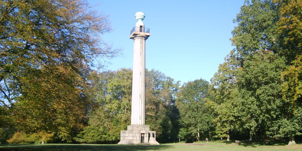 A tall stone column with a statue on top, surrounded by green trees and clear blue skies, situated in a park setting.