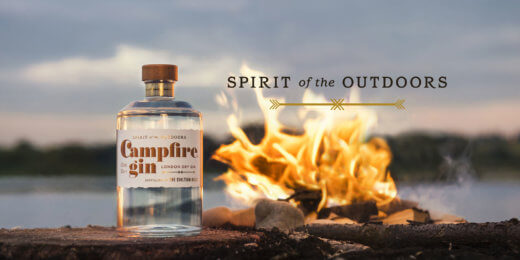 A bottle of Campfire gin is placed on a log near a glowing campfire, with a serene outdoor background. The image captures a cozy, adventurous spirit of the outdoors.