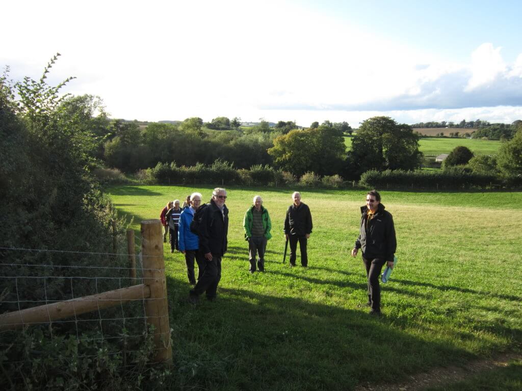 A group of six people, dressed for outdoor activity, walking along a path in a green, open field with trees and a cloudy sky in the background.