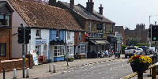 A quaint street view featuring a row of charming shops with varied facades, surrounded by trees and traffic lights. A few vehicles are parked along the road, creating a lively atmosphere in this picturesque neighborhood.