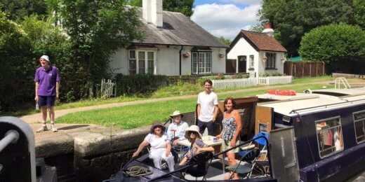 A group of people, including children, enjoy a sunny day on a narrowboat along a canal, with a residential house in the background and a person standing on the bank.