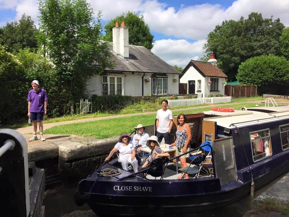 A group of people, including children, enjoy a sunny day on a narrowboat along a canal, with a residential house in the background and a person standing on the bank.