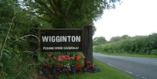 A roadside sign for the village of Wigginton, surrounded by greenery and flowers, with a message encouraging cautious driving.