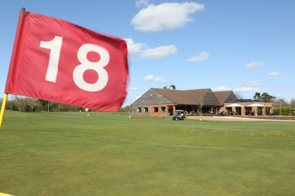 A close-up of a red golf flag marked "18," with a clubhouse and green grass in the background under a blue sky.