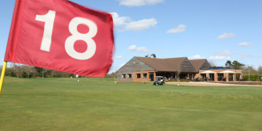 A close-up of a red golf flag marked "18," with a clubhouse and green grass in the background under a blue sky.