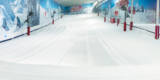 An indoor skiing facility featuring a smooth, snow-covered slope and scenic mountain murals on the walls. The environment is bright and spacious, designed for skiing or snowboarding activities.