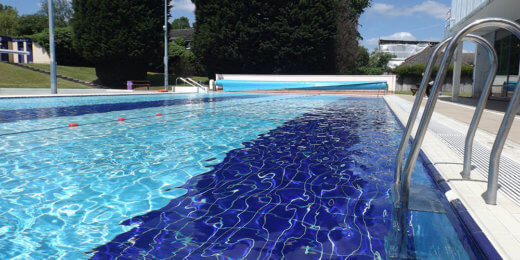 A swimming pool features shimmering blue water reflecting the sky, with a white tiled edge and metal ladder. Lush green trees line the background, indicating a sunny day, creating a refreshing and inviting atmosphere.