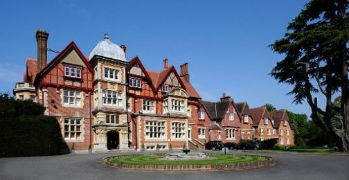 A picturesque, historic mansion featuring a mix of red brick and stone architecture, surrounded by lush greenery and a circular driveway. The building showcases ornate details and is set against a clear blue sky, creating an inviting and serene atmosphere.
