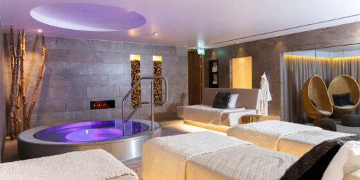 A modern spa interior featuring relaxing massage tables, a circular hot tub with ambient lighting, and comfortable seating areas. The space exudes tranquility, inviting guests to unwind and rejuvenate.
