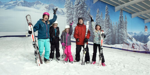 A group of five children and adults stands on a snowy slope, smiling and posing with skis in hand. They are in an indoor ski facility, with a mountain mural in the background, dressed in colorful winter gear.