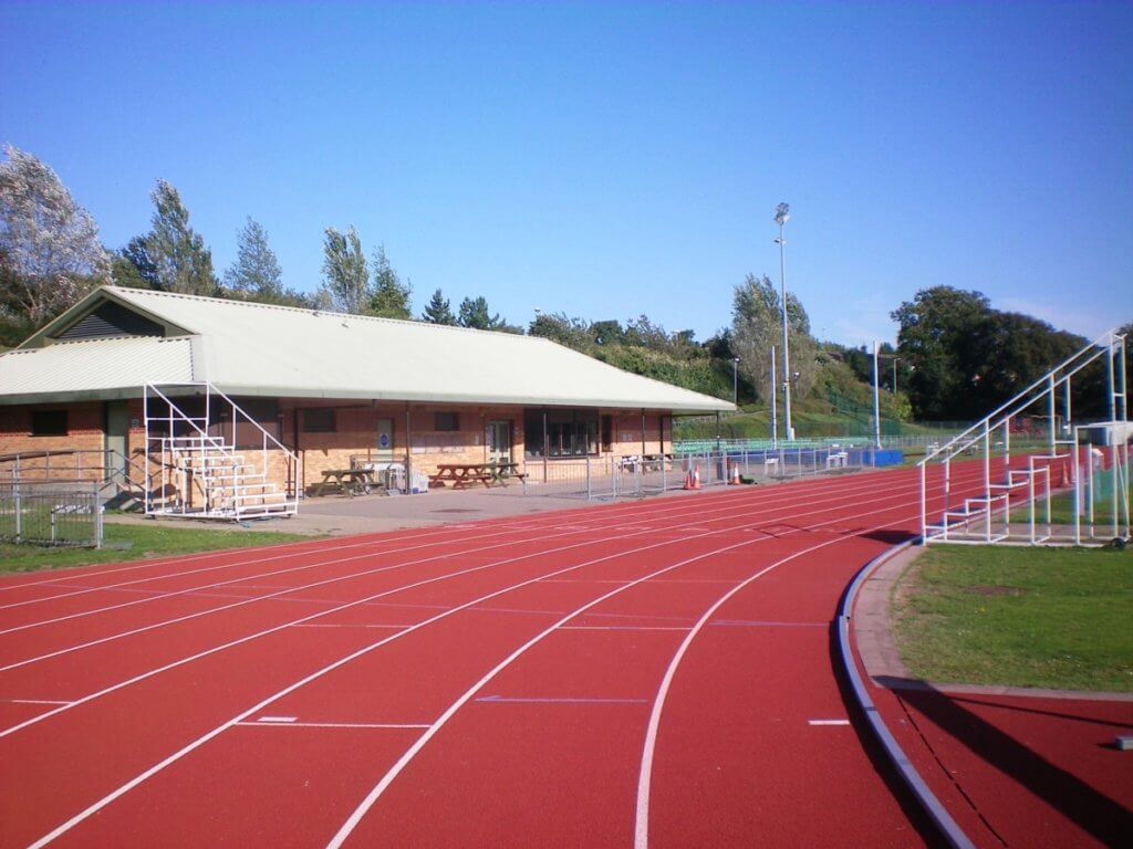 A red athletic track curves in the foreground, leading to a brick building with a green roof. Surrounded by trees and fields, the area appears sunny and open, ideal for sports and activities.