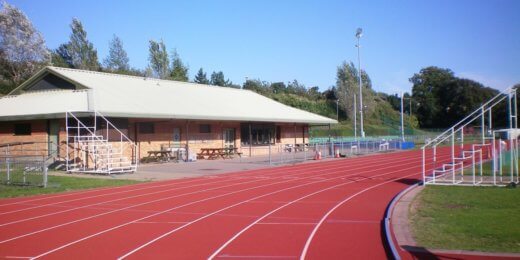 A red athletic track curves in the foreground, leading to a brick building with a green roof. Surrounded by trees and fields, the area appears sunny and open, ideal for sports and activities.
