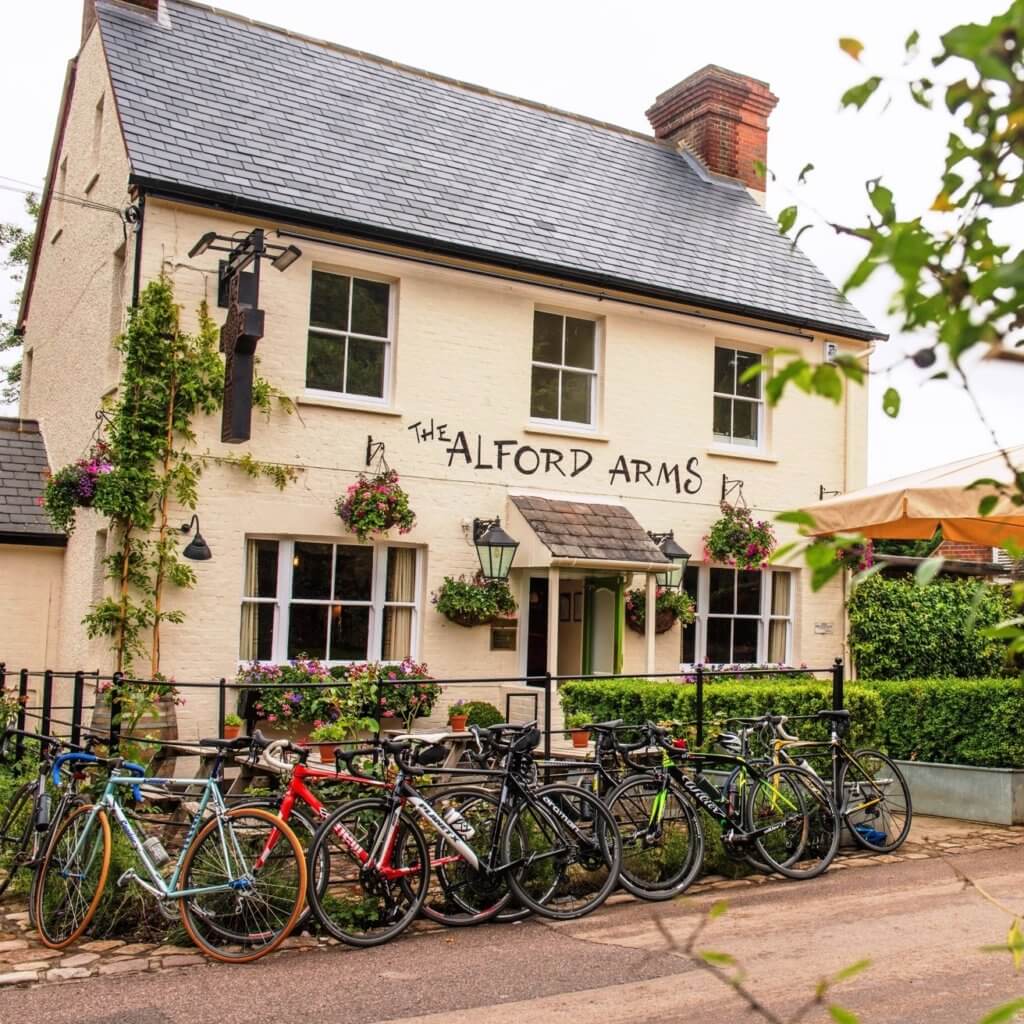 A quaint pub named "The Alford Arms," featuring a charming, rustic exterior adorned with plants. Bicycles are parked in front, highlighting a welcoming atmosphere for cyclists and visitors.