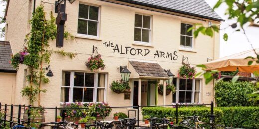 A quaint pub named "The Alford Arms," featuring a charming, rustic exterior adorned with plants. Bicycles are parked in front, highlighting a welcoming atmosphere for cyclists and visitors.
