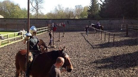 A group of individuals engaged in horseback riding practice in an enclosed arena. One rider in armor holds a spear, while others are riding or walking with horses, surrounded by a gravel surface and trees in the background.