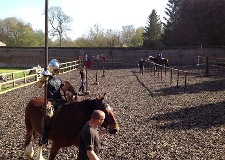 A group of individuals engaged in horseback riding practice in an enclosed arena. One rider in armor holds a spear, while others are riding or walking with horses, surrounded by a gravel surface and trees in the background.
