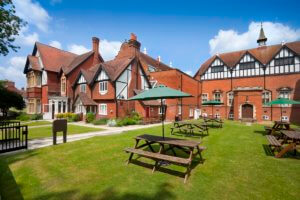 A picturesque view of a red-brick building with a classic architectural style, surrounded by green lawns and picnic tables. The outdoor area features umbrellas for shade, set against a clear blue sky with fluffy white clouds.