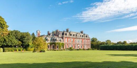 A grand historic mansion with red brick exterior is set against a clear blue sky and lush green lawn, surrounded by trees.